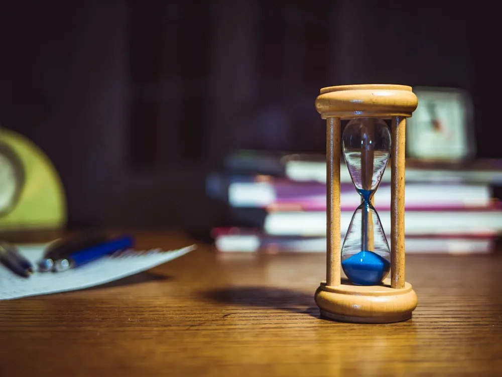 A wooden hourglass on a desk beside writing materials, books, and analog clocks.